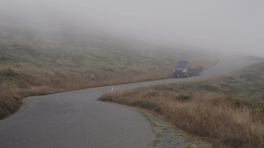The video shows a winding asphalt road cutting through dry grassy hillsides that are heavily shrouded in a thick atmospheric foga darkcolored mercedesbenz sprinterstyle van emerges from the mist and drives down the road toward the camera emphasiz