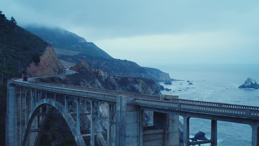 On a moody overcast day a dark van drives across a large concrete bridge spanning a rugged and foggy coastline