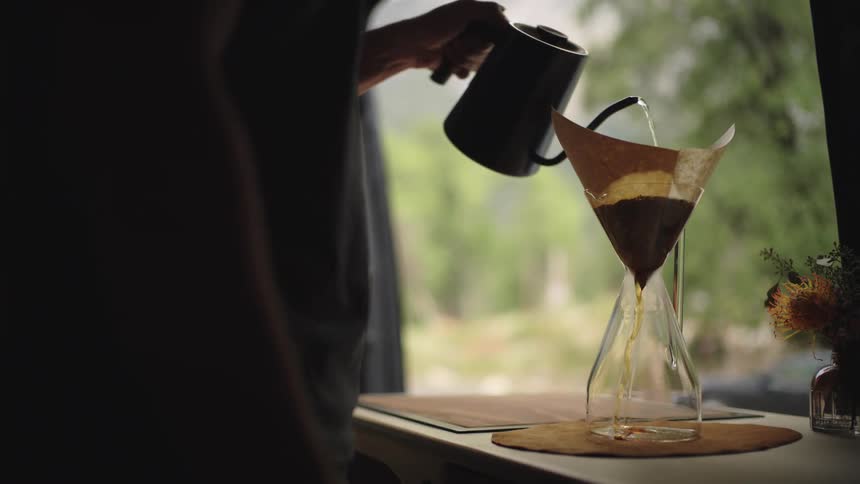 A person holding a kettle with hot water and pouring the water into a glass jar to brew coffee through a paper filter
