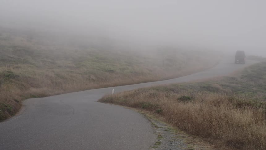 A dark van drives around a bend in a remote road on a foggy day