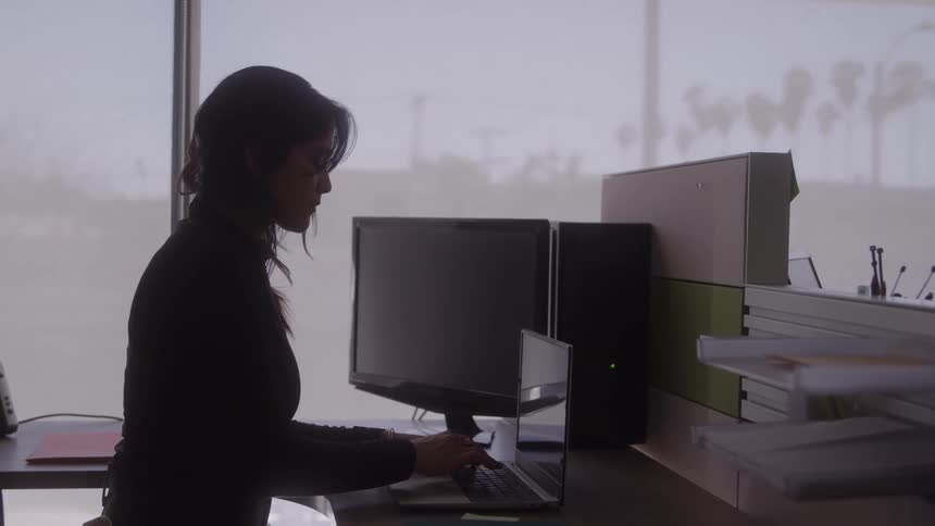 A woman is sitting at a desk in an office typing on a laptop