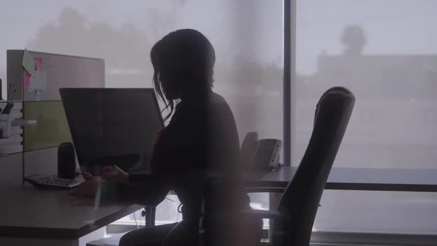 A figure is shown in silhouette working at a desktop computer in a modern office illuminated primarily by a large window behind them the person is seated in an office chair and appears to be typing or interacting with the screen