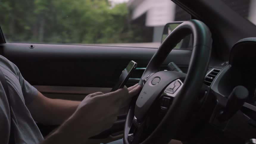 The video is a closeup shot inside a car showing the drivers hands and the steering wheelthe driver is holding a smartphone with his left hand and actively using it with his right index finger while appearing to be in motion or stopped briefly on