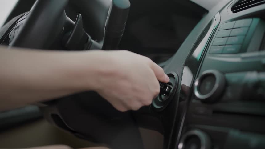 A hand inserts a metal key into a vehicle ignition cylinder the key rotates clockwise to engage the starter and the key then returns to the auxiliary position all within the dark interior of a car cabin
