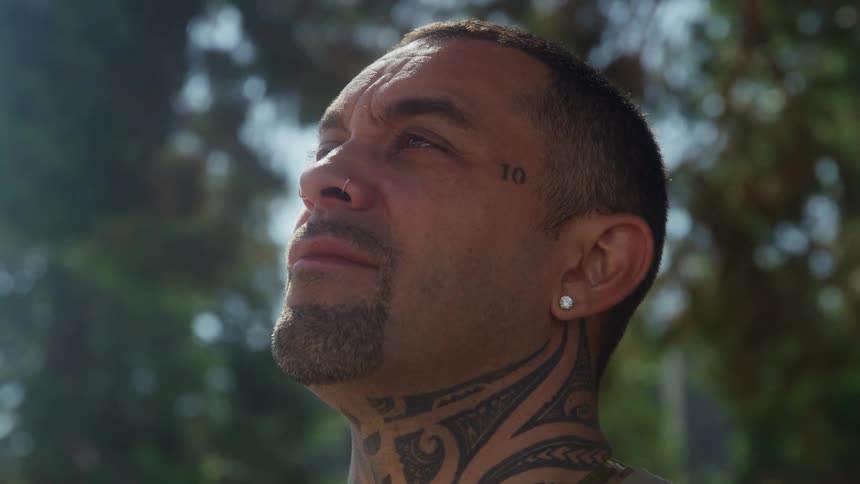 A man with extensive neck tattoos stands outdoors looks upward and bright sunlight shines from above through blurred foliage in the background