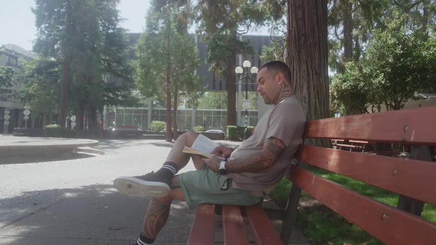 A man sits on a wooden bench reading a book in a sunny urban park