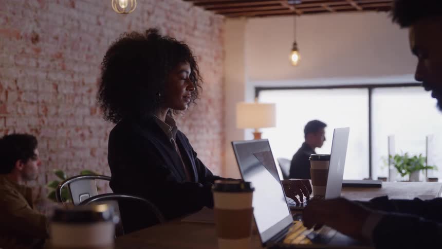 Young black woman wearing a suit typing on a laptop computer over a wooden desk surrounded by other people doing similar activities in an office indoors during day time