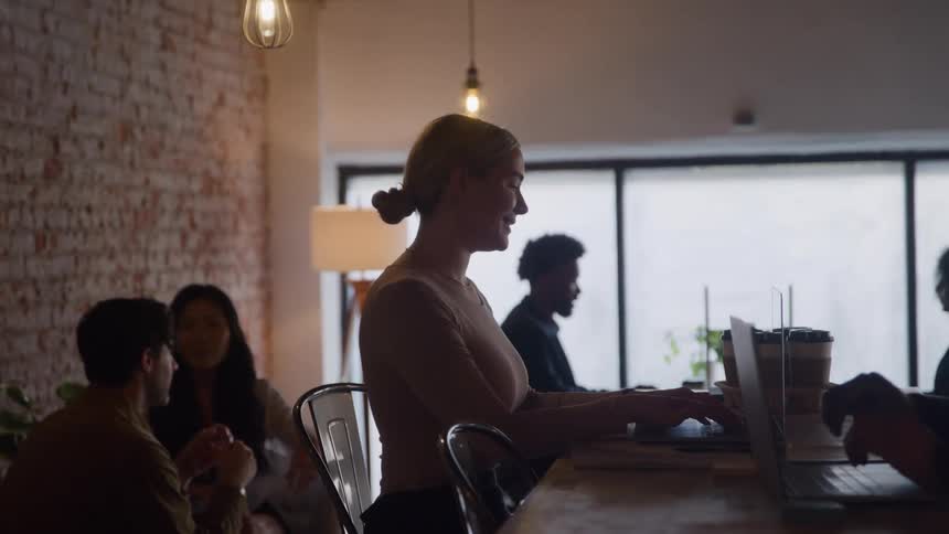 Young white woman sitting down and typing on a laptop computer whilte talking to other people around her in an office space indoors during day time
