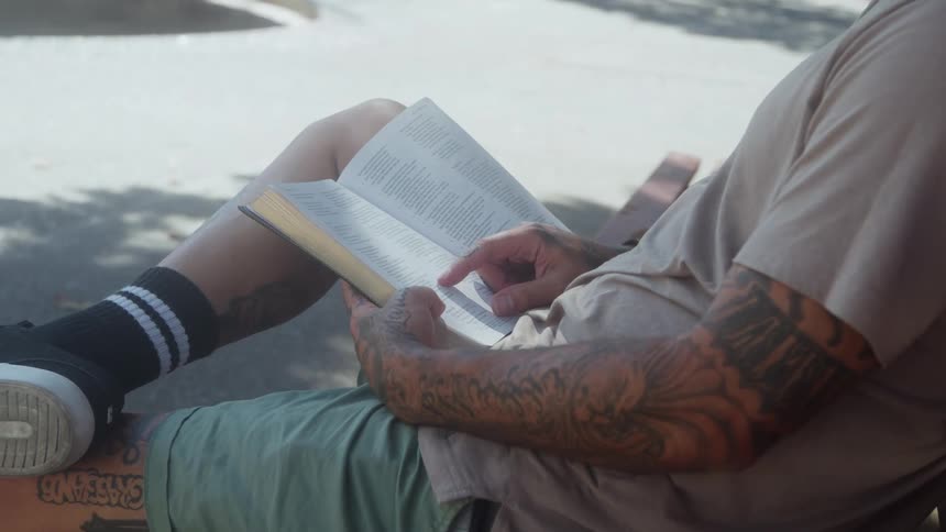 A man sits on a bench outdoors reading a book in the sunlight