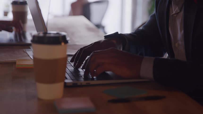 Hands of a man wearing a suit typing on a laptop computer over a wooden desk in an office space with two more people around indoors during day time