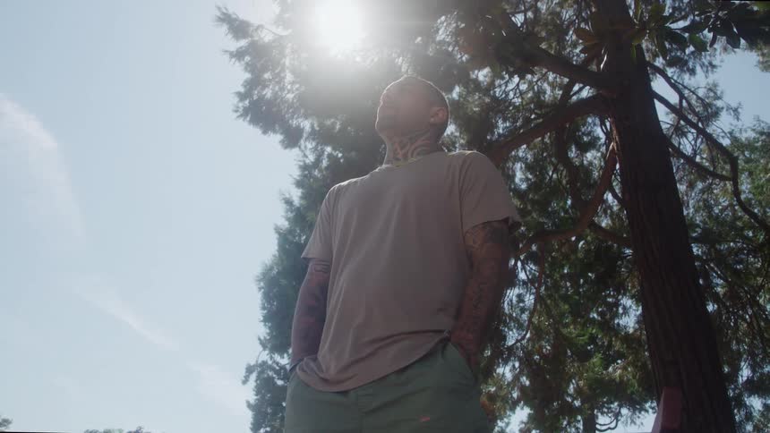 The video shows a lowangle shot of a man standing outdoors wearing a tshirt and shorts with a tattoo visible on his neck and armsthe bright sunlight is positioned directly behind his head creating a strong silhouette and lens flare an