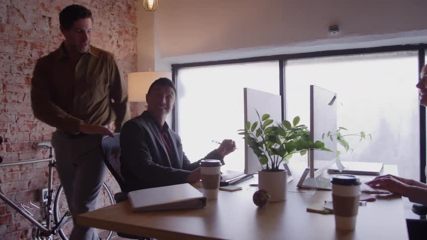 An office with two workers a man and a woman sitting at a desk working on a computer while another man walks by communicating with them at midday