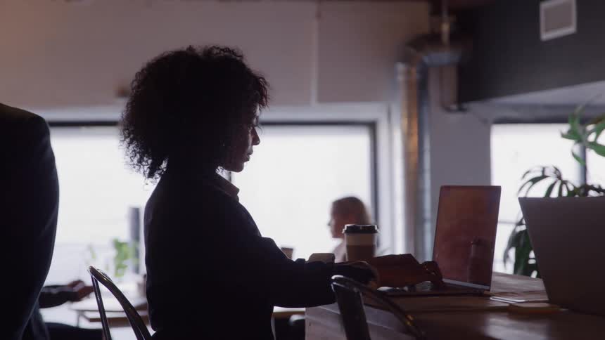 A woman sitting in a caf with a laptop on the table typing on it a man walks past her and in the background a man and a woman are sitting