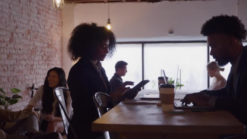 Two people sit at a table in the foreground one uses a laptop and the other uses a tablet while several other people sit or stand in the background all within an indoor area with a brick wall and bright ambient lighting