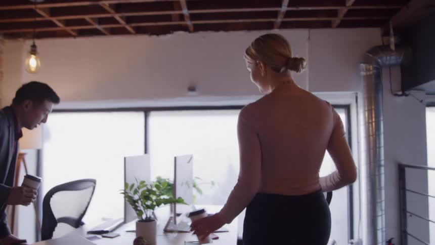 Blonde young woman walking and carrying a beverage on her hand then sitting down in front of a computer in a desk in an office surrounded by other coworkers indoors during day time
