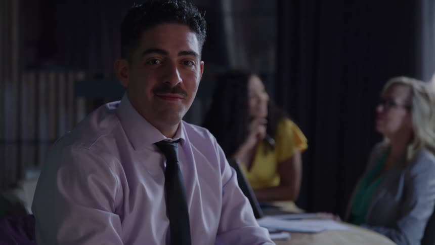 A young man wearing a tie sits in a conference room at midday while two women sit behind him engaged in a discussion