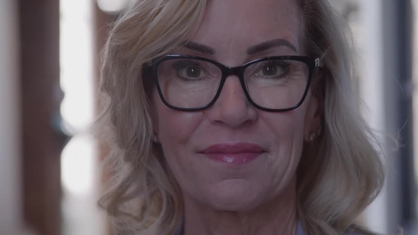 A woman with blonde hair and black glasses stands in a welllit indoor area she makes direct eye contact with the camera and smiles the background is blurred