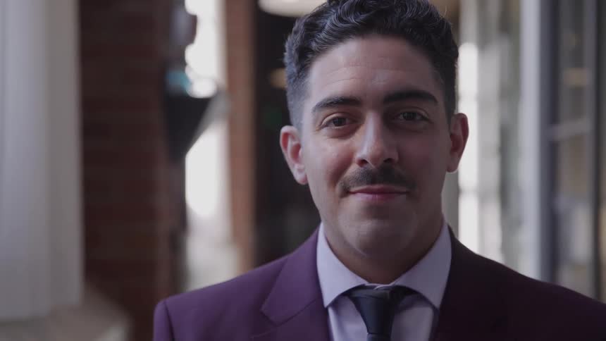 The video is a mediumcloseup of a man in a maroon suit jacket and dark tie looking offcamera then turning to face the camerahe has dark slickedback hair and a mustache appearing to be indoors in a formal or professional setting