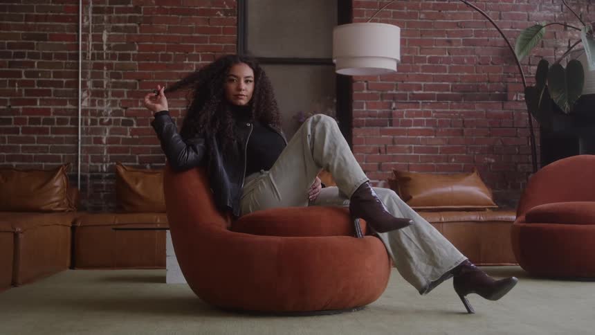 A young woman sits on a sofa wearing a black jacket jeans and highheeled shoes rubbing her hands through her long curly hair