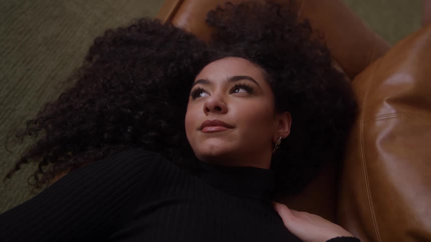 A close view shows a woman with long dark curly hair wearing a black sweater lying down on a brown leather couch and looking thoughtfully to the side