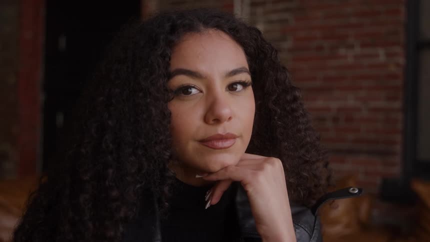 A close view of a young woman with long dark curly hair shows her smiling while resting her chin on her hand wearing a black leather jacket against a blurred brick background