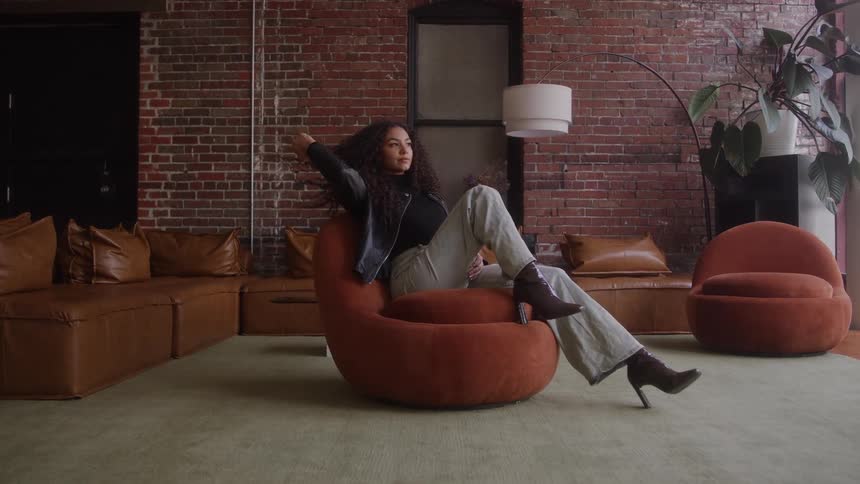 The video features a stylish woman sitting casually in a curved rustcolored armchair in a loftstyle roomshe is dressed in a black top leather jacket and lightwash jeans with dark highheeled boots against a backdrop of exposed brick wa