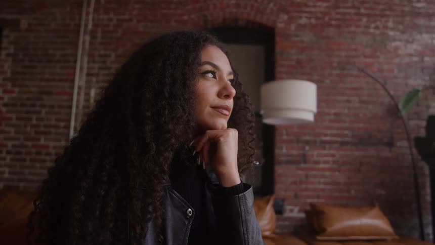 Woman in a black jacket looks thoughtful in a room with brick walls