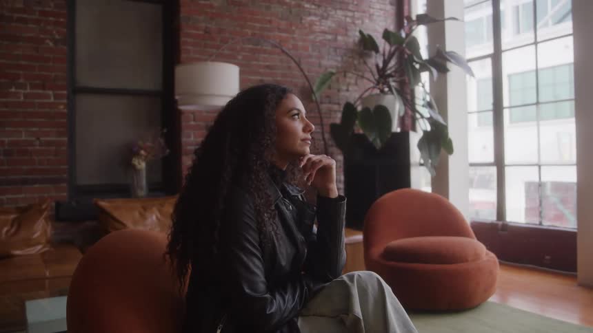 A woman with long curly hair is sitting on an orange chair in a brightly lit modern loft wearing a leather jacket and looking thoughtfully offcamera the apartment features exposed brick walls and large windows suggesting an urban converted industria