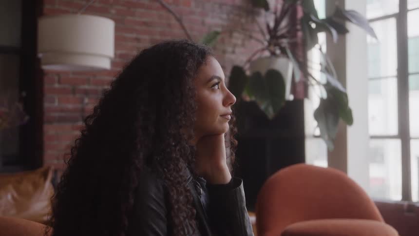 A young woman with long curly hair sits indoors alone lost in deep sad thought