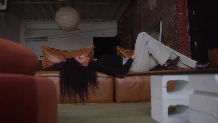 The video is a wideangle shot of a woman with long curly hair lying back on a brown leather couch in a loftstyle roomshe is dressed in lightcolored pants and boots with one leg bent and she appears to be relaxing in a casual moody setting