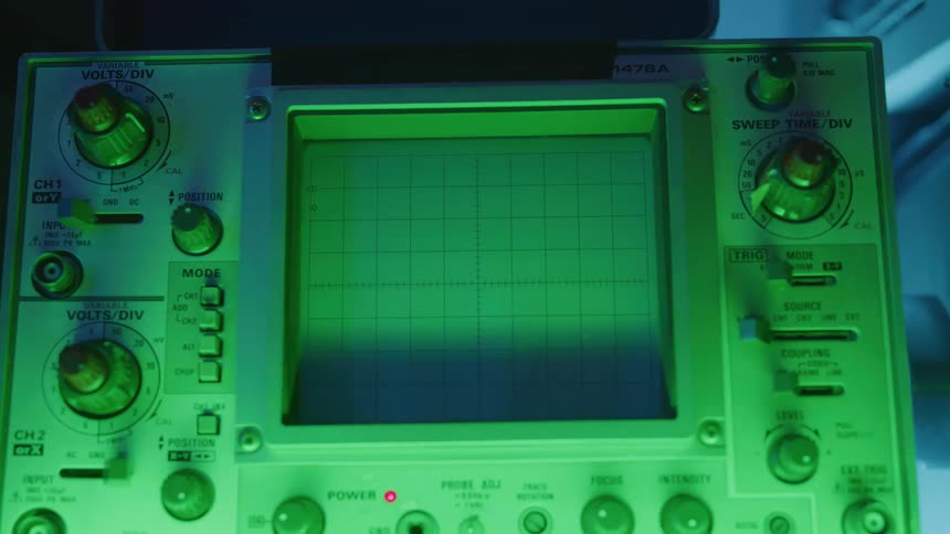 A close view of an old oscilloscope machine shows its square green gridded screen and all its dials and controls illuminated by a soft green and blue light