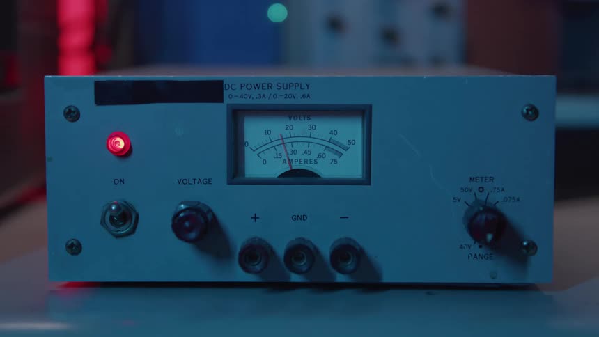 A close view of an old dc power supply machine shows a hand flipping the power switch on causing a red indicator light and the analog volts and amperes meter to illuminate under cool blue lighting