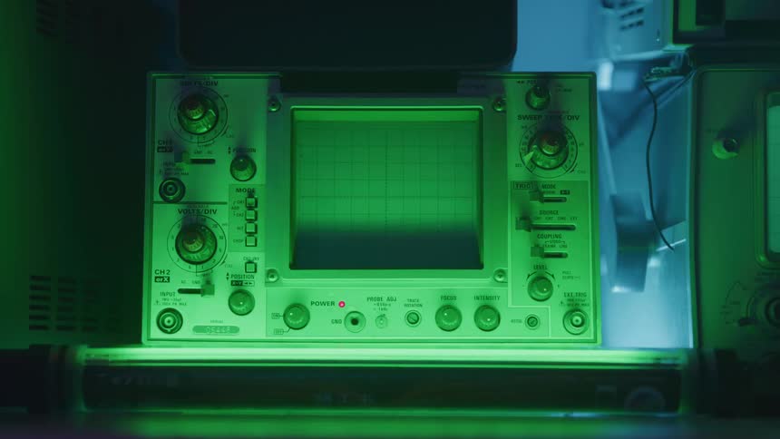 An analog oscilloscope sits on a surface illuminated by green light in a dark room