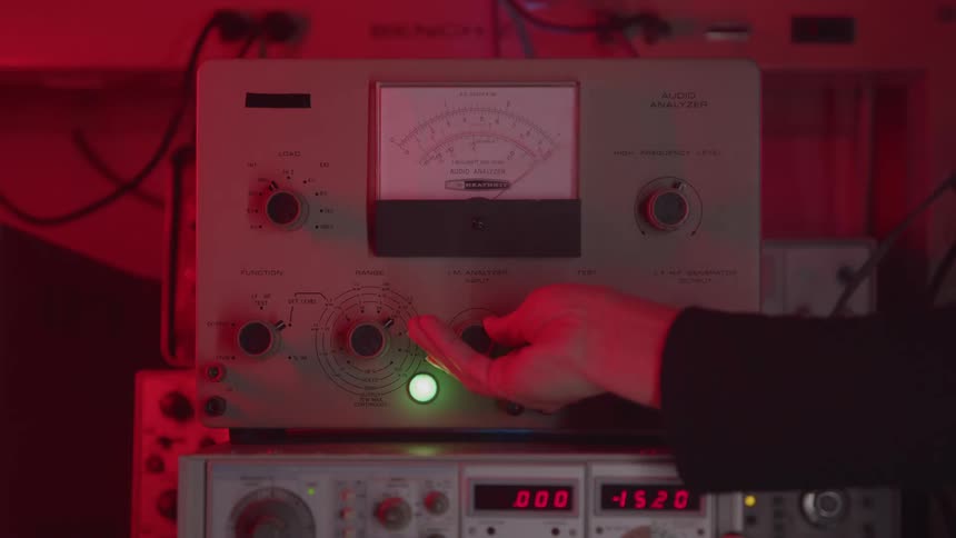 A close view of an audio analyzer machine shows a hand reaching in to adjust a control knob on the front panel with a circular gauge under dramatic red ambient light