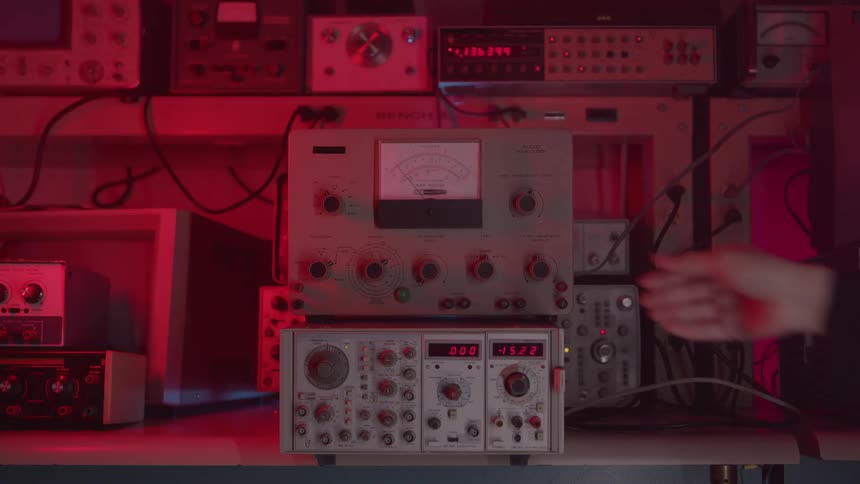 The video shows a closeup of a hand in a dark sleeve adjusting a knob on a piece of vintage electronic lab equipment the background is filled with various other pieces of testing equipment and is bathed in a red glow