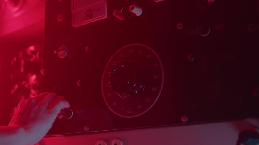 The video is a closeup of a persons hand illuminated by a deep red light operating an analog machine control panelthe hand turns a circular knob on the complex interface suggesting a technical or industrial setting like a darkroom or lab