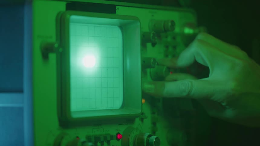 Hand operating the knobs of a greenlit oscilloscope