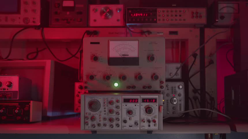 A hand reaches into the frame to turn a knob on a piece of vintage analog testing equipment the equipment sits on a surface surrounded by stacks of other similar devices and the entire scene is illuminated with a strong red light cast over the background