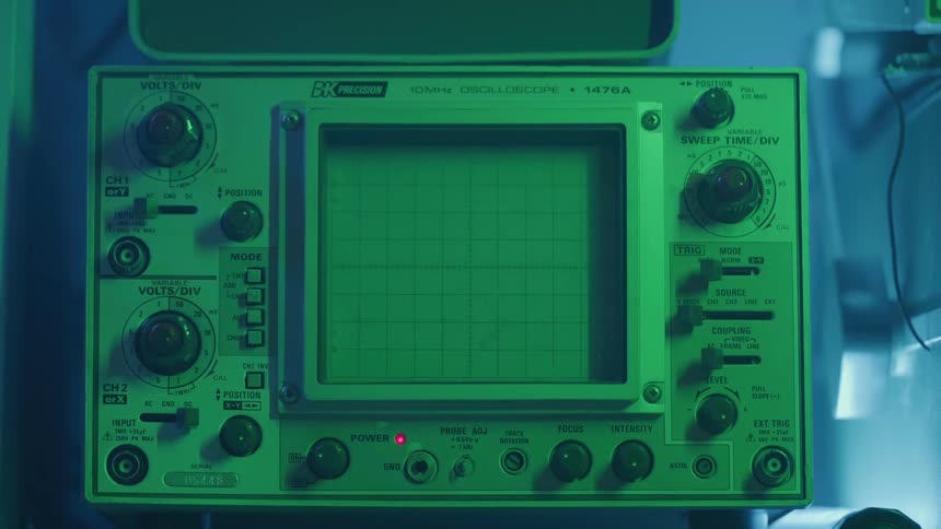 A persons hand adjusts an analog oscilloscope under greenblue lighting