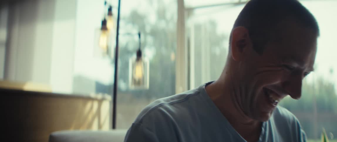 A laughing bald man in a light blue tshirt raises both fists in a gesture of success and excitement while sitting near a bright window indoors