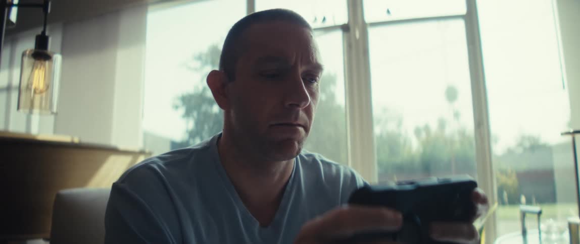 A man sits indoors and holds a mobile video game with both hands he looks intently at the screen while moving his thumbs and the scene is brightly lit by natural light coming through a large window behind him