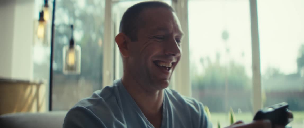 White man wearing a blue shirt typing on a smartphone and laughing indoors during day time