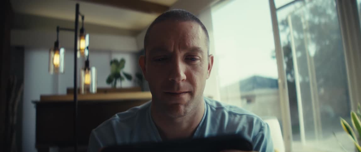 A man with a shaved head sits indoors and holds a mobile phone with both hands he stares intently at the screen and bright natural light illuminates his face from a large window on the right side of the frame