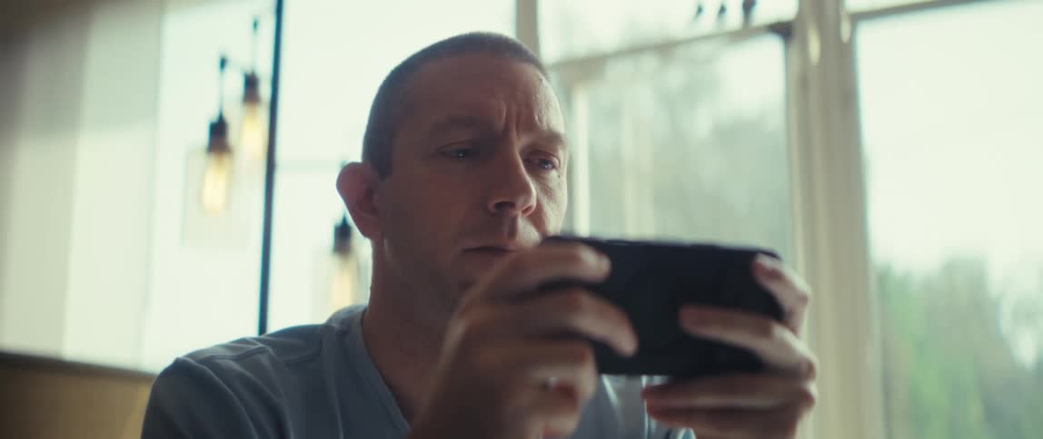 A man with short hair sits indoors and looks down with concentration at a black smartphone he holds horizontally his face is illuminated by bright light from a large window behind him