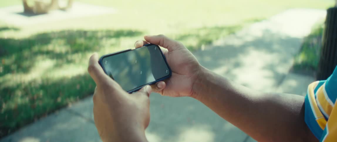 A closeup shot focuses on a persons hands holding a smartphone horizontally ready to capture or view content the person who appears to be wearing a blue and yellow jersey is outdoors in a sunny park setting with grass visible in the background