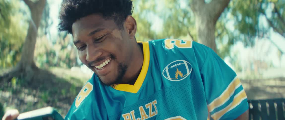 The video is a mediumclose shot of a man wearing a blue and yellow sports jersey with the number 22 sitting outdoors and looking at his smartphonehe is smiling broadly and laughing as he watches the screen suggesting he is finding something hi