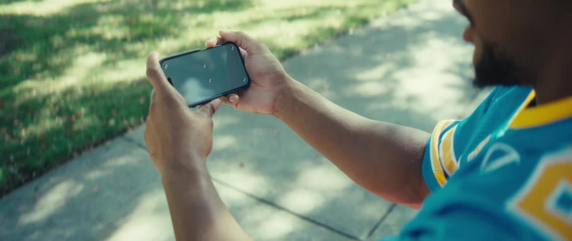 Man wears a jersey while sitting on a park bench and cheering as he watches his smartphone