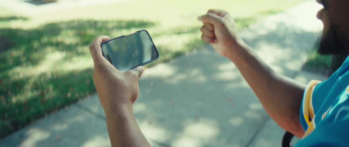 A young man sits on a public bench under a tree watching a game on his mobile device and celebrating a goal scored in the game