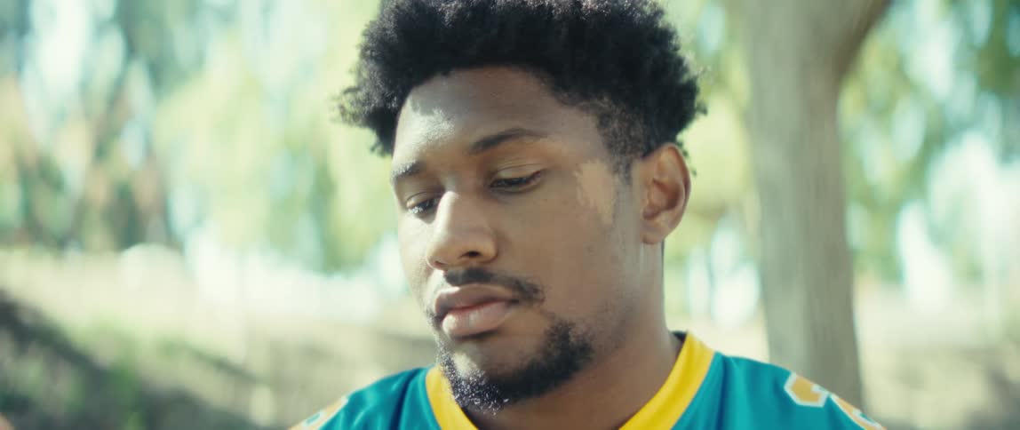 A young black man wearing a jersey sits on a bench in a public park watching content on his mobile phone under a tree at midday