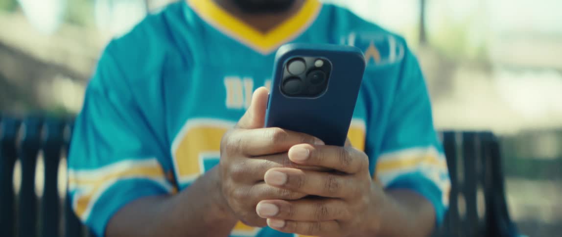 A young man wearing a blue jersey sits on a bench in a public park at midday holding an iphone
